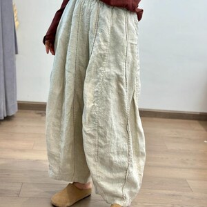 May include: Light beige linen pants with vertical seam details. The pants have a wide, relaxed fit and are gathered at the waist. The model is wearing a maroon long-sleeve shirt and tan suede slippers.