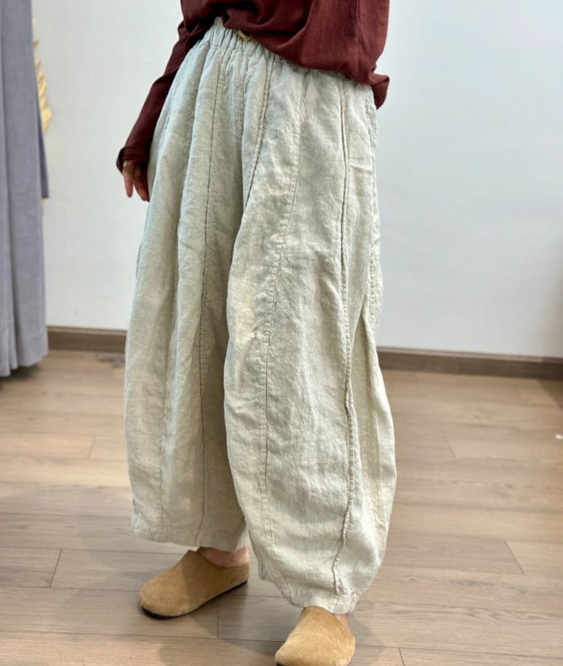 May include: Light beige linen pants with vertical seam details. The pants have a wide, relaxed fit and are gathered at the waist. The model is wearing a maroon long-sleeve shirt and tan suede slippers.