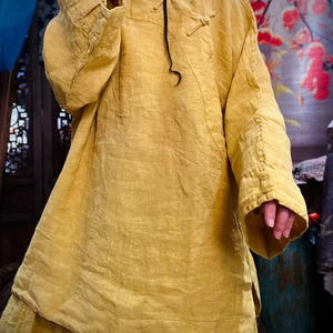 May include: A loose-fitting, long-sleeved, yellow linen tunic with a mandarin collar and a knotted closure at the neckline.