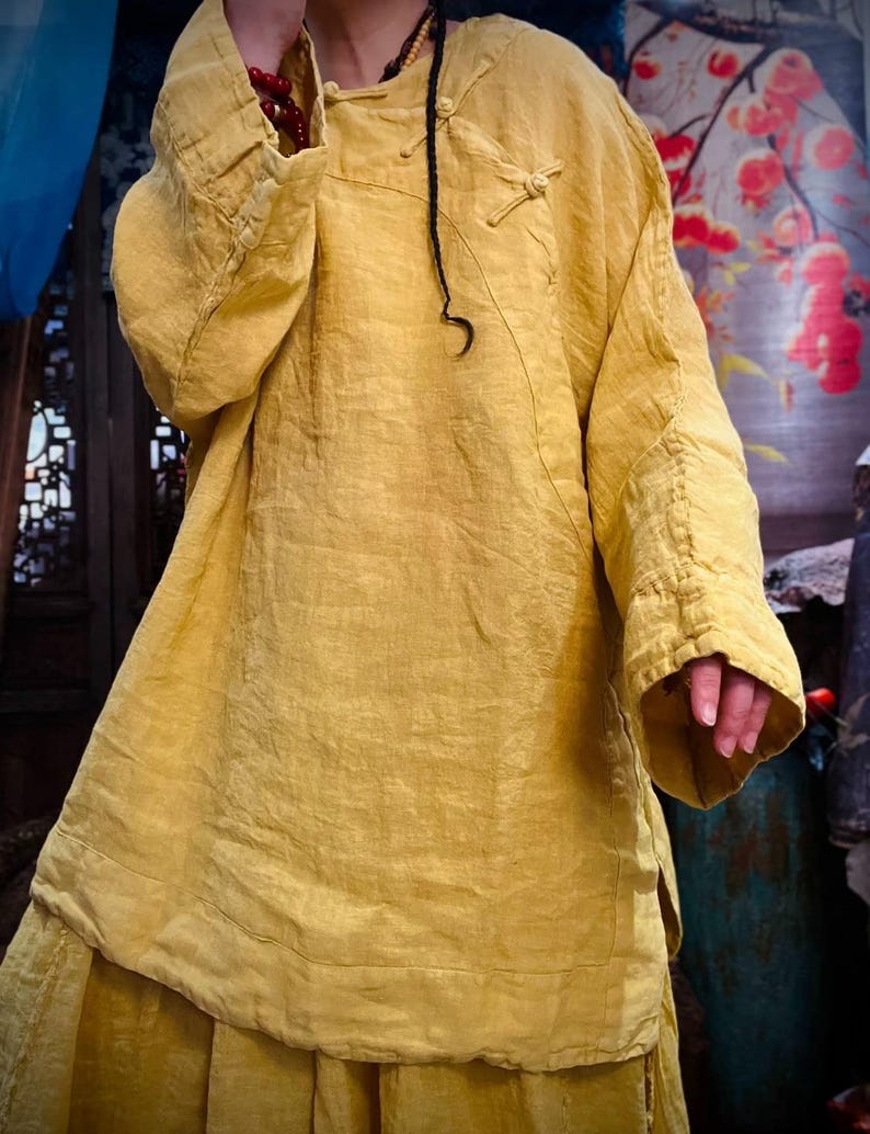 May include: A loose-fitting, long-sleeved, yellow linen tunic with a mandarin collar and a knotted closure at the neckline.