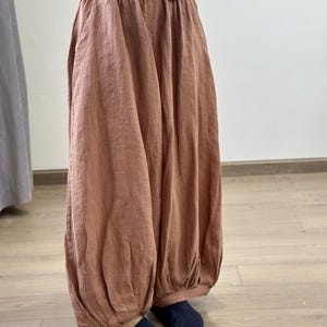 May include: Loose-fitting, rust-colored linen pants with a gathered waist and ankle cuffs. The pants have a relaxed, flowing silhouette, suggesting comfort and casual wear. The fabric appears lightweight and breathable, ideal for warmer weather.