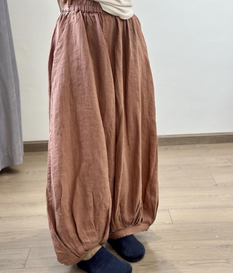 May include: Loose-fitting, rust-colored linen pants with a gathered waist and ankle cuffs. The pants have a relaxed, flowing silhouette, suggesting comfort and casual wear. The fabric appears lightweight and breathable, ideal for warmer weather.