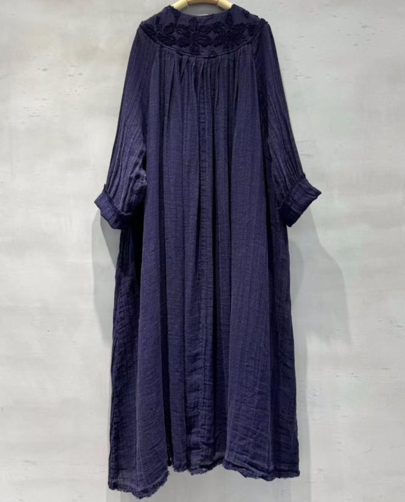May include: A long, navy blue dress with a floral embroidered neckline. The dress features long sleeves with rolled cuffs and a loose, flowing silhouette. The fabric appears to be a lightweight, textured material.