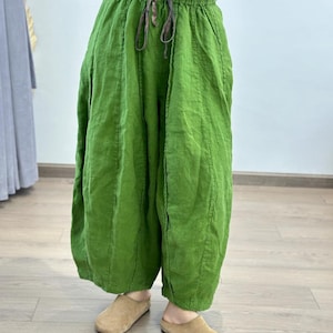 May include: Bright green linen pants with a drawstring waist. The pants have a loose, flowing design and are paired with tan suede slip-on shoes. The fabric appears to be slightly wrinkled, giving a casual, comfortable look. The pants are a vibrant, solid color.