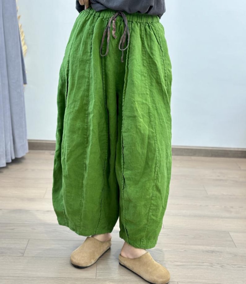 May include: Bright green linen pants with a drawstring waist. The pants have a loose, flowing design and are paired with tan suede slip-on shoes. The fabric appears to be slightly wrinkled, giving a casual, comfortable look. The pants are a vibrant, solid color.