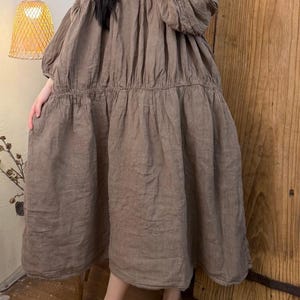 May include: A brown linen dress with a V-neck and long, gathered sleeves. The dress has a tiered skirt and a relaxed fit, suitable for casual wear. The dress is knee-length and appears to be made of a lightweight fabric.