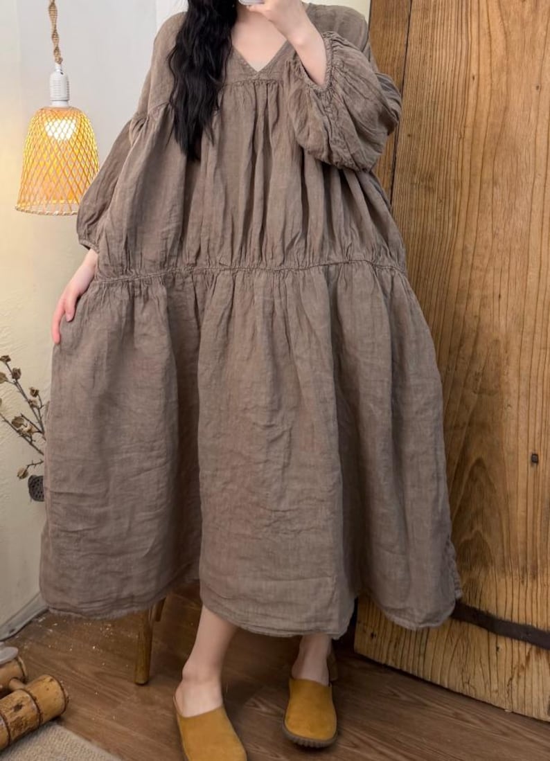 May include: A brown linen dress with a V-neck and long, gathered sleeves. The dress has a tiered skirt and a relaxed fit, suitable for casual wear. The dress is knee-length and appears to be made of a lightweight fabric.