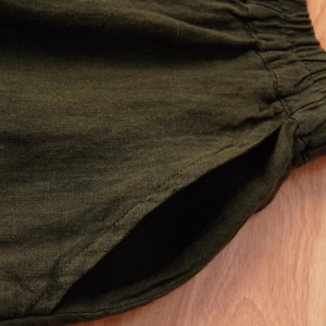 May include: Close-up of olive green linen shorts with a visible pocket and elastic waistband. The fabric has a textured appearance, suggesting a breathable and comfortable material. The shorts are likely for casual wear.