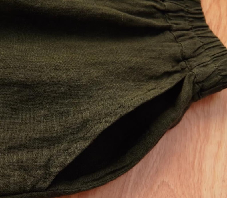 May include: Close-up of olive green linen shorts with a visible pocket and elastic waistband. The fabric has a textured appearance, suggesting a breathable and comfortable material. The shorts are likely for casual wear.
