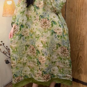 May include: A long, flowing dress with a floral pattern in shades of green, beige, and pink. The dress has long sleeves and a round neckline with green trim. The hemline and sleeve cuffs are also trimmed in green. The dress is made of a lightweight fabric.