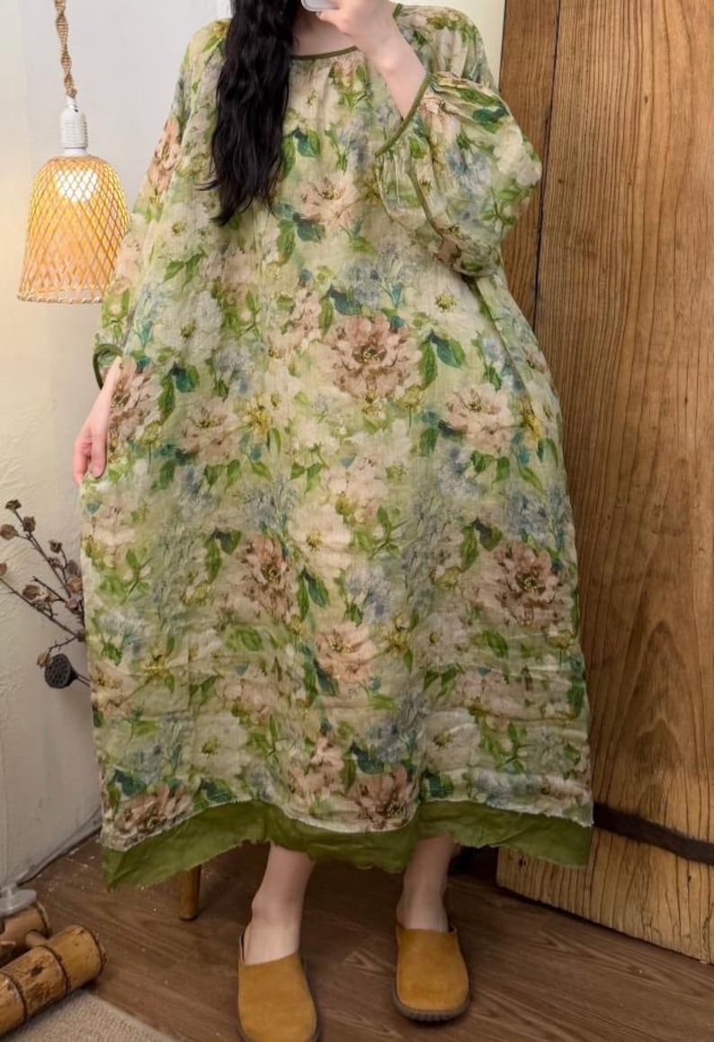 May include: A long, flowing dress with a floral pattern in shades of green, beige, and pink. The dress has long sleeves and a round neckline with green trim. The hemline and sleeve cuffs are also trimmed in green. The dress is made of a lightweight fabric.