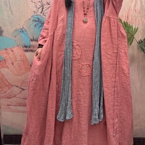 May include: A long, flowing coral dress with long sleeves and decorative patches. A grey scarf is draped around the neck. The dress appears to be made of a soft, lightweight fabric, suitable for casual wear.