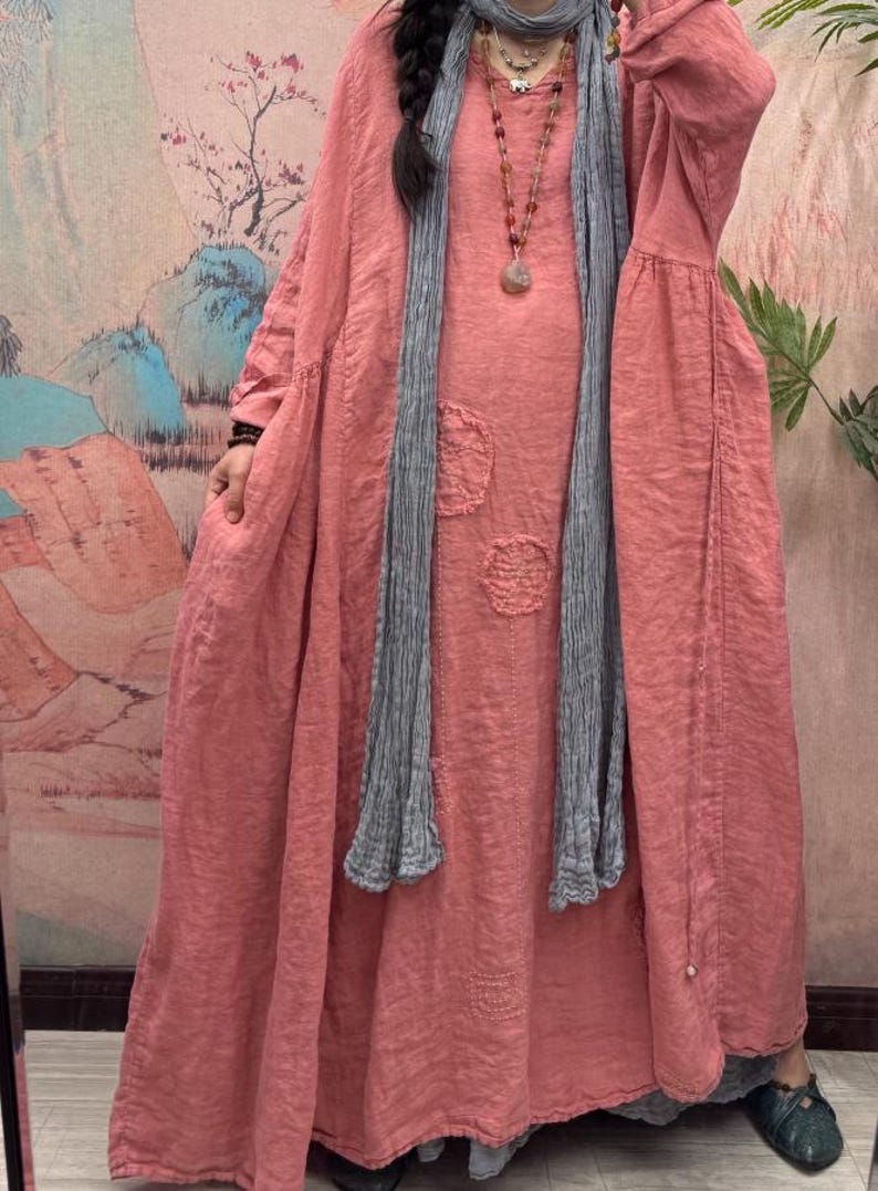 May include: A long, flowing coral dress with long sleeves and decorative patches. A grey scarf is draped around the neck. The dress appears to be made of a soft, lightweight fabric, suitable for casual wear.