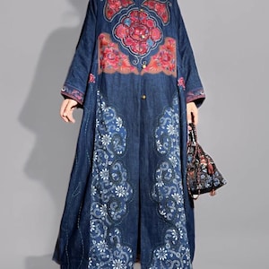 May include: A long, dark blue denim coat with intricate floral embroidery in red, pink, and white. The coat features a mandarin collar, long sleeves, and a full-length design. A small, patterned handbag is held in the hand.