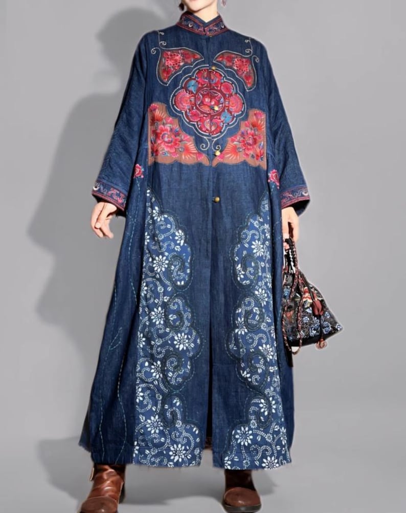 May include: A long, dark blue denim coat with intricate floral embroidery in red, pink, and white. The coat features a mandarin collar, long sleeves, and a full-length design. A small, patterned handbag is held in the hand.