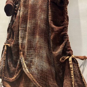 May include: A brown, textured garment with long sleeves and a cinched waist. The fabric has a subtle pattern and is trimmed with a lighter brown material. The garment features ties at the waist and hem, adding a decorative element.