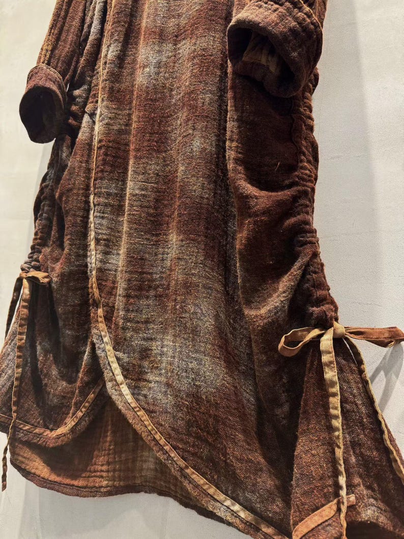 May include: A brown, textured garment with long sleeves and a cinched waist. The fabric has a subtle pattern and is trimmed with a lighter brown material. The garment features ties at the waist and hem, adding a decorative element.