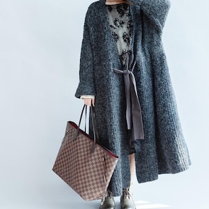 May include: A long, grey, textured coat with a matching tie belt. The coat is worn over a floral print dress. A large brown and tan checked tote bag is held in the left hand. The boots are grey.
