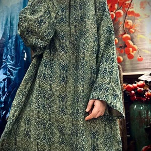 May include: A loose-fitting, long-sleeved tunic featuring an intricate, repeating damask-style pattern in shades of forest green, olive, and dark blue. The garment has a round neckline and wide, flowing sleeves that extend past the wrists.