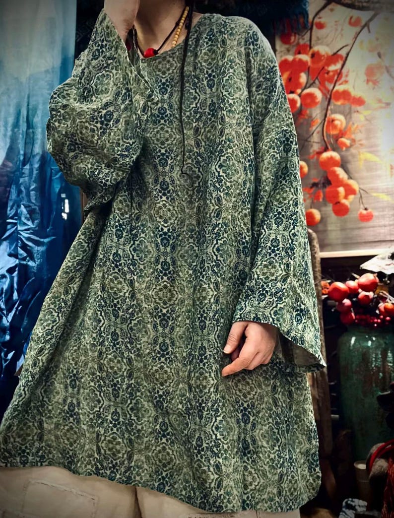 May include: A loose-fitting, long-sleeved tunic featuring an intricate, repeating damask-style pattern in shades of forest green, olive, and dark blue. The garment has a round neckline and wide, flowing sleeves that extend past the wrists.
