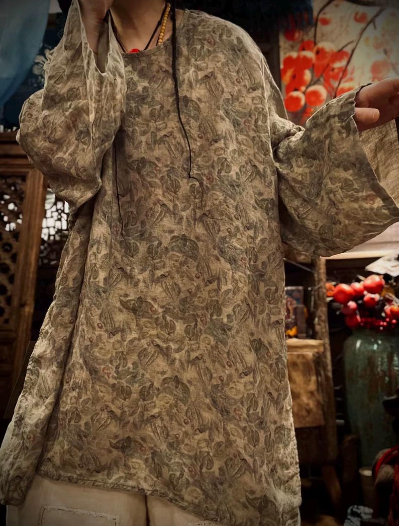 May include: A long-sleeved tunic featuring a muted earth-toned botanical print with leaves and small berries in shades of sage green, brown, and beige. The garment has a round neckline, a loose, relaxed fit, and wide sleeves that extend past the wrists.