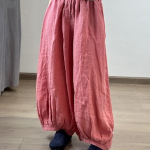 May include: Loose-fitting, coral-colored linen pants with a gathered waist and elasticized ankles. The pants have a relaxed, flowing silhouette. The model is wearing navy blue shoes. The pants are made of a lightweight fabric.