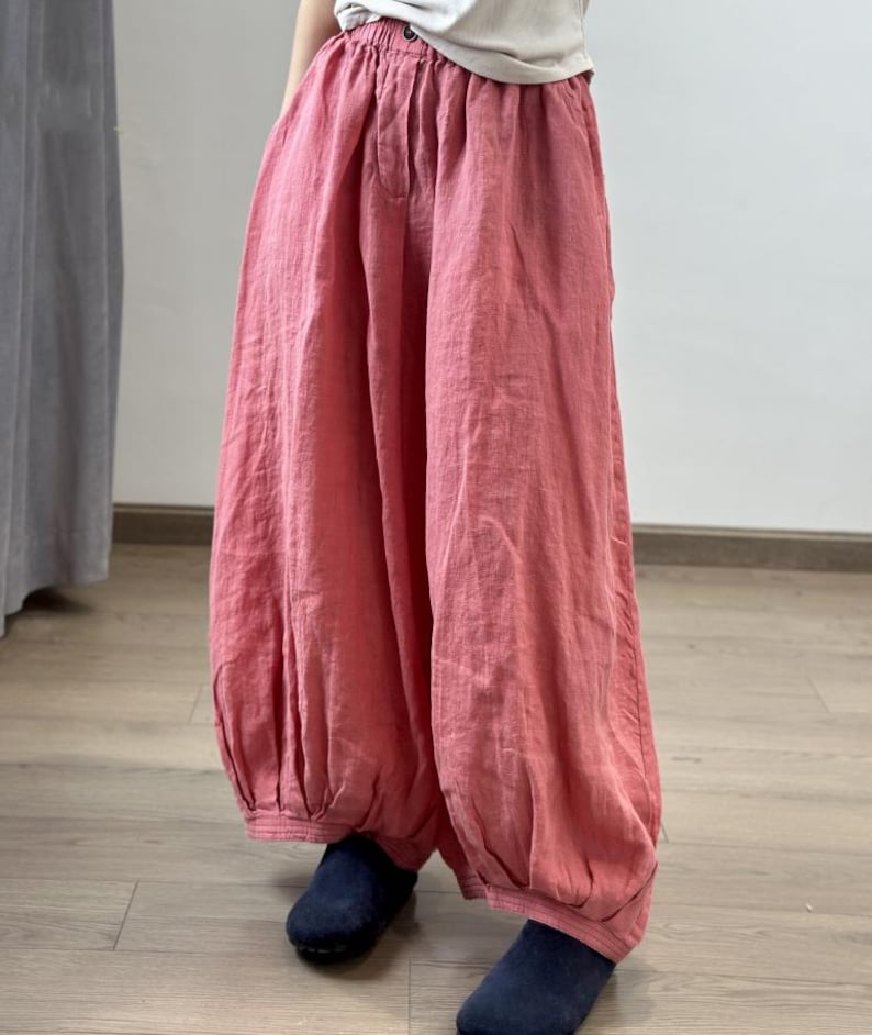 May include: Loose-fitting, coral-colored linen pants with a gathered waist and elasticized ankles. The pants have a relaxed, flowing silhouette. The model is wearing navy blue shoes. The pants are made of a lightweight fabric.