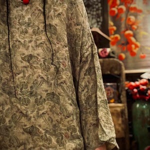 May include: A long-sleeved shirt featuring an all-over botanical print in muted shades of olive green, brown, and tan. The garment has a round neckline and a loose, relaxed fit. A black cord necklace with a single red bead is worn around the neck.