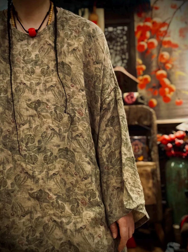 May include: A long-sleeved shirt featuring an all-over botanical print in muted shades of olive green, brown, and tan. The garment has a round neckline and a loose, relaxed fit. A black cord necklace with a single red bead is worn around the neck.