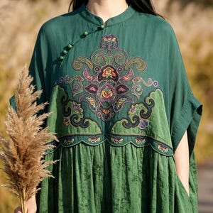 May include: A dark green dress featuring a mandarin collar with small spherical buttons along the diagonal neckline. The bodice displays an intricate, multi-colored floral embroidery with scalloped edges, while the skirt has a subtle texture.