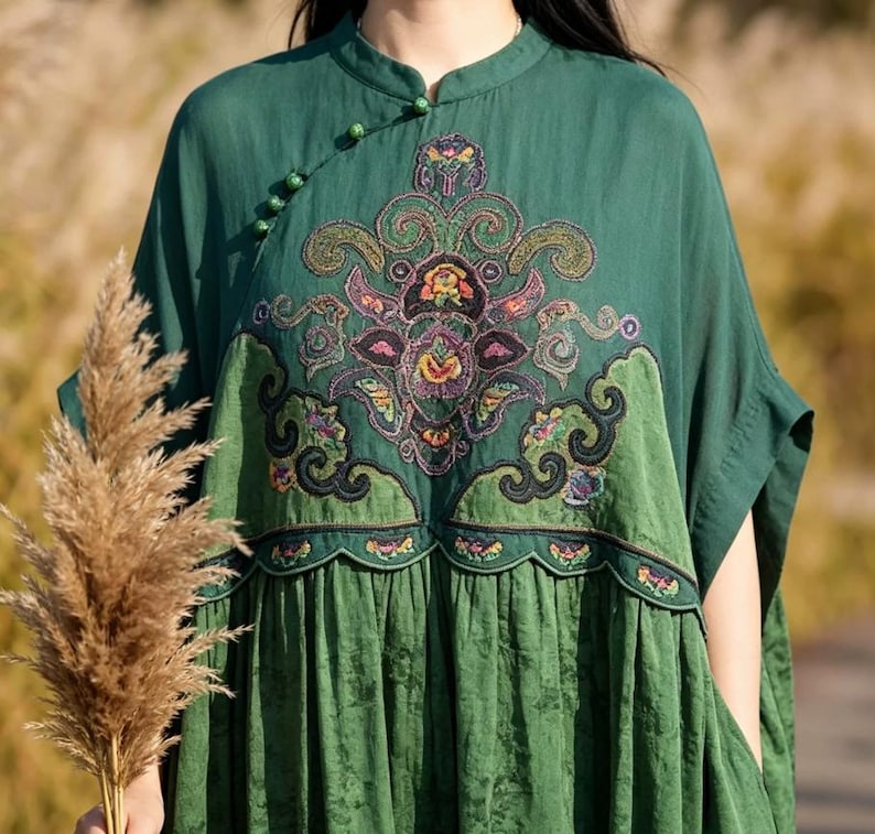 May include: A dark green dress featuring a mandarin collar with small spherical buttons along the diagonal neckline. The bodice displays an intricate, multi-colored floral embroidery with scalloped edges, while the skirt has a subtle texture.