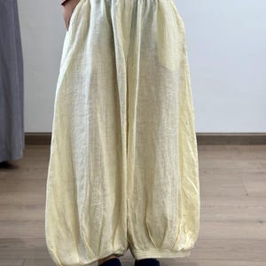 May include: Pale yellow linen pants with a gathered waist and balloon-style legs. The pants have a relaxed fit and pockets. The fabric appears lightweight and breathable, suitable for warm weather. The pants are paired with dark blue socks.