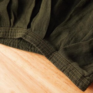 May include: Close-up of olive green fabric with visible texture and stitching details. The fabric appears to be gathered at the waistline, with a contrasting gold-colored thread used for the stitching. The garment is likely a skirt or pants.