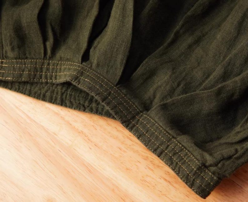 May include: Close-up of olive green fabric with visible texture and stitching details. The fabric appears to be gathered at the waistline, with a contrasting gold-colored thread used for the stitching. The garment is likely a skirt or pants.