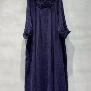 May include: A long, navy blue dress with long sleeves and a V-neckline. The neckline features intricate dark embroidery. The dress has a loose, flowing silhouette and a textured fabric. The sleeves have a cuff detail, and the hem is slightly frayed.