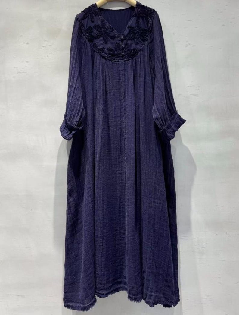 May include: A long, navy blue dress with long sleeves and a V-neckline. The neckline features intricate dark embroidery. The dress has a loose, flowing silhouette and a textured fabric. The sleeves have a cuff detail, and the hem is slightly frayed.