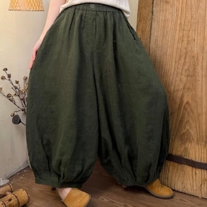 May include: Dark green linen pants with a gathered waist and voluminous, balloon-like legs. The pants are paired with tan shoes and a light-colored top. The fabric appears soft and flowing, suggesting a comfortable fit. The style is casual and relaxed.