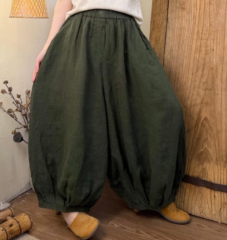 May include: Dark green linen pants with a gathered waist and voluminous, balloon-like legs. The pants are paired with tan shoes and a light-colored top. The fabric appears soft and flowing, suggesting a comfortable fit. The style is casual and relaxed.