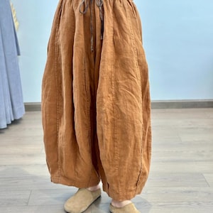 May include: Rust-colored linen pants with a drawstring waist and wide, gathered legs. The pants have a relaxed fit and are paired with tan suede slip-on shoes. The fabric appears to be lightweight and breathable, suitable for casual wear.