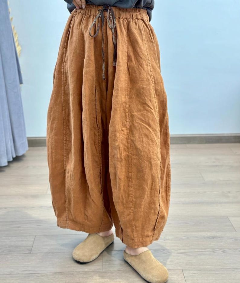 May include: Rust-colored linen pants with a drawstring waist and wide, gathered legs. The pants have a relaxed fit and are paired with tan suede slip-on shoes. The fabric appears to be lightweight and breathable, suitable for casual wear.