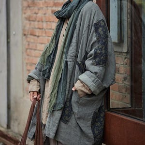 May include: A long, oversized gray coat with patches of patterned fabric and a matching scarf. The coat has large pockets and a relaxed fit, suitable for layering. The outfit is completed with a brown tote bag.