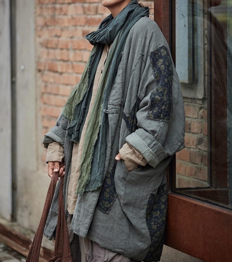 May include: A long, oversized gray coat with patches of patterned fabric and a matching scarf. The coat has large pockets and a relaxed fit, suitable for layering. The outfit is completed with a brown tote bag.