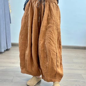 May include: Wide-leg, rust-colored linen pants with a drawstring waist. The pants have a relaxed fit and are made of a textured fabric. The pants are paired with beige slip-on shoes. The pants are a casual style.