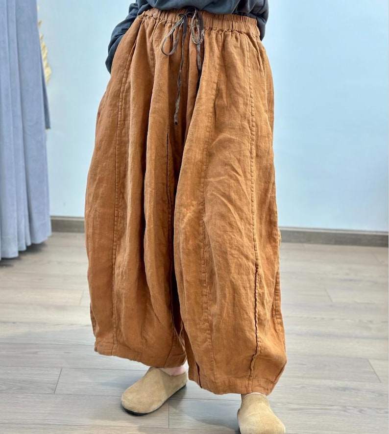 May include: Wide-leg, rust-colored linen pants with a drawstring waist. The pants have a relaxed fit and are made of a textured fabric. The pants are paired with beige slip-on shoes. The pants are a casual style.