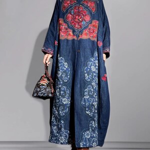 May include: A long, dark blue denim coat with intricate floral embroidery in red, pink, and white. The coat features a mandarin collar and a button-down front. A small, patterned handbag complements the outfit. The coat's design is reminiscent of traditional Asian styles.