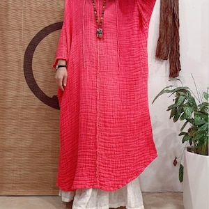 May include: A long, pink, loose-fitting dress with a V-neck and 3/4 sleeves. The dress is made of a textured fabric and has a relaxed fit.