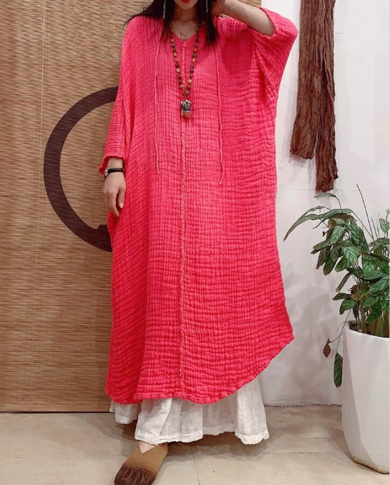 May include: A long, pink, loose-fitting dress with a V-neck and 3/4 sleeves. The dress is made of a textured fabric and has a relaxed fit.