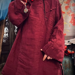 May include: A burgundy linen tunic with long sleeves and a mandarin collar. The tunic has a button closure at the neck and a decorative knot detail on the front.