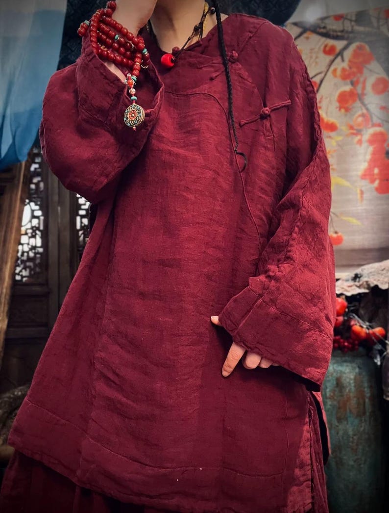 May include: A burgundy linen tunic with long sleeves and a mandarin collar. The tunic has a button closure at the neck and a decorative knot detail on the front.