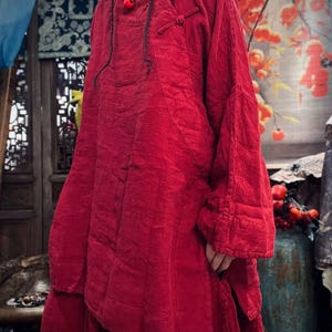 May include: A loose-fitting, long-sleeved, red tunic top with a mandarin collar and a knotted closure at the neckline.